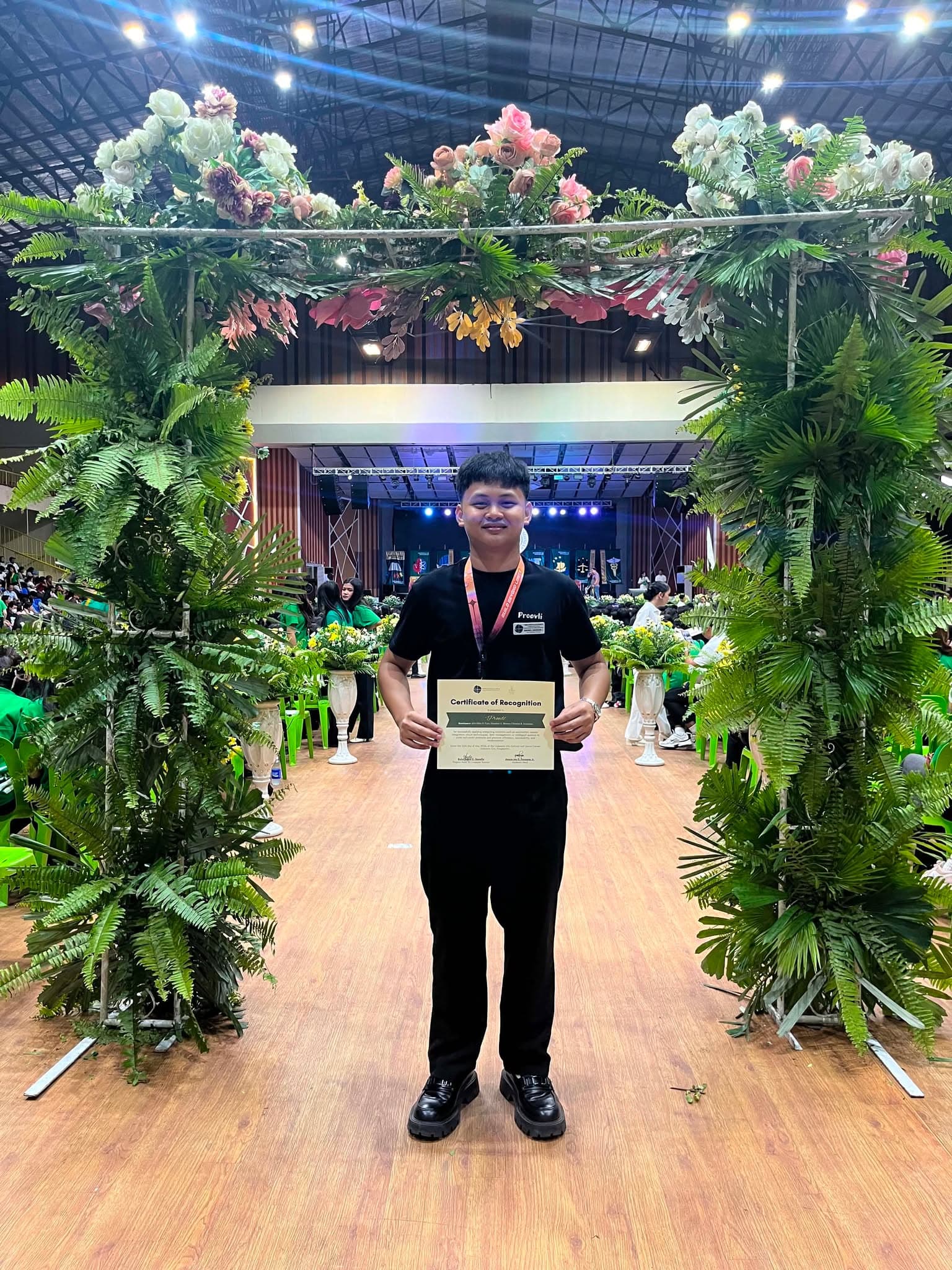 A Proovli team member posing with the certificate of recognition beneath the floral stage arch.